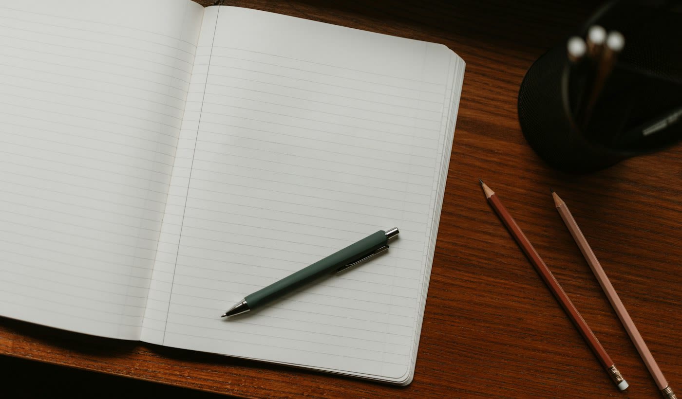 An open blank notebook with a pen and pencils on a dark wooden desk