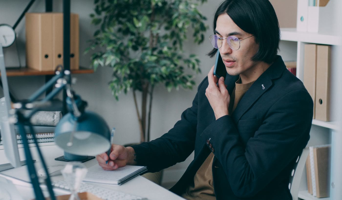 A man on a phone call writing notes by hand at his desk