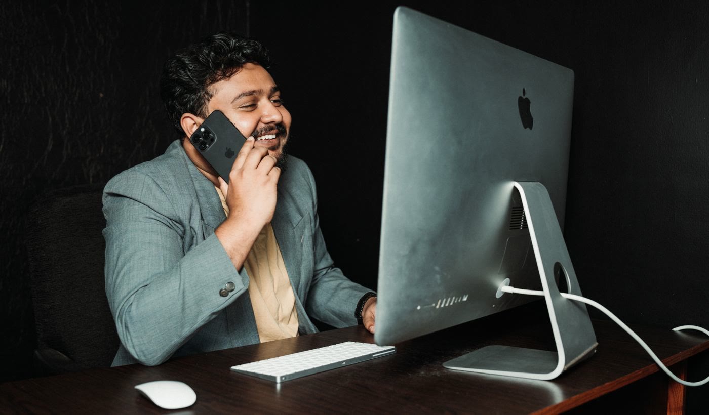 A man in a blazer smiling on a phone call at his office desk